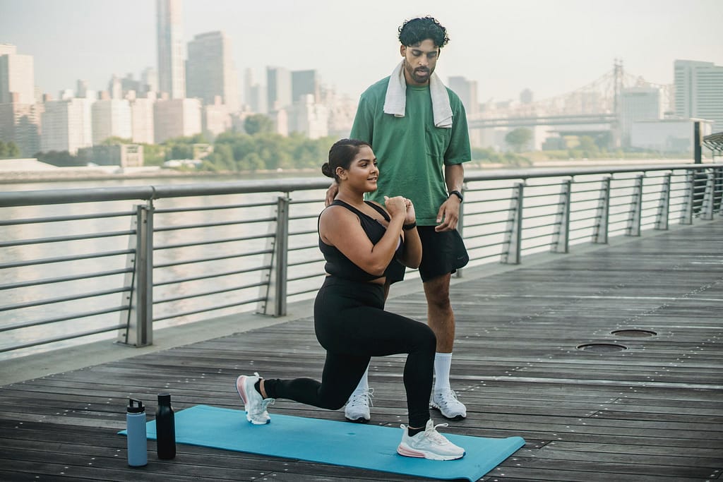 Woman performing lunges on a boardwalk with a trainer by the river view.